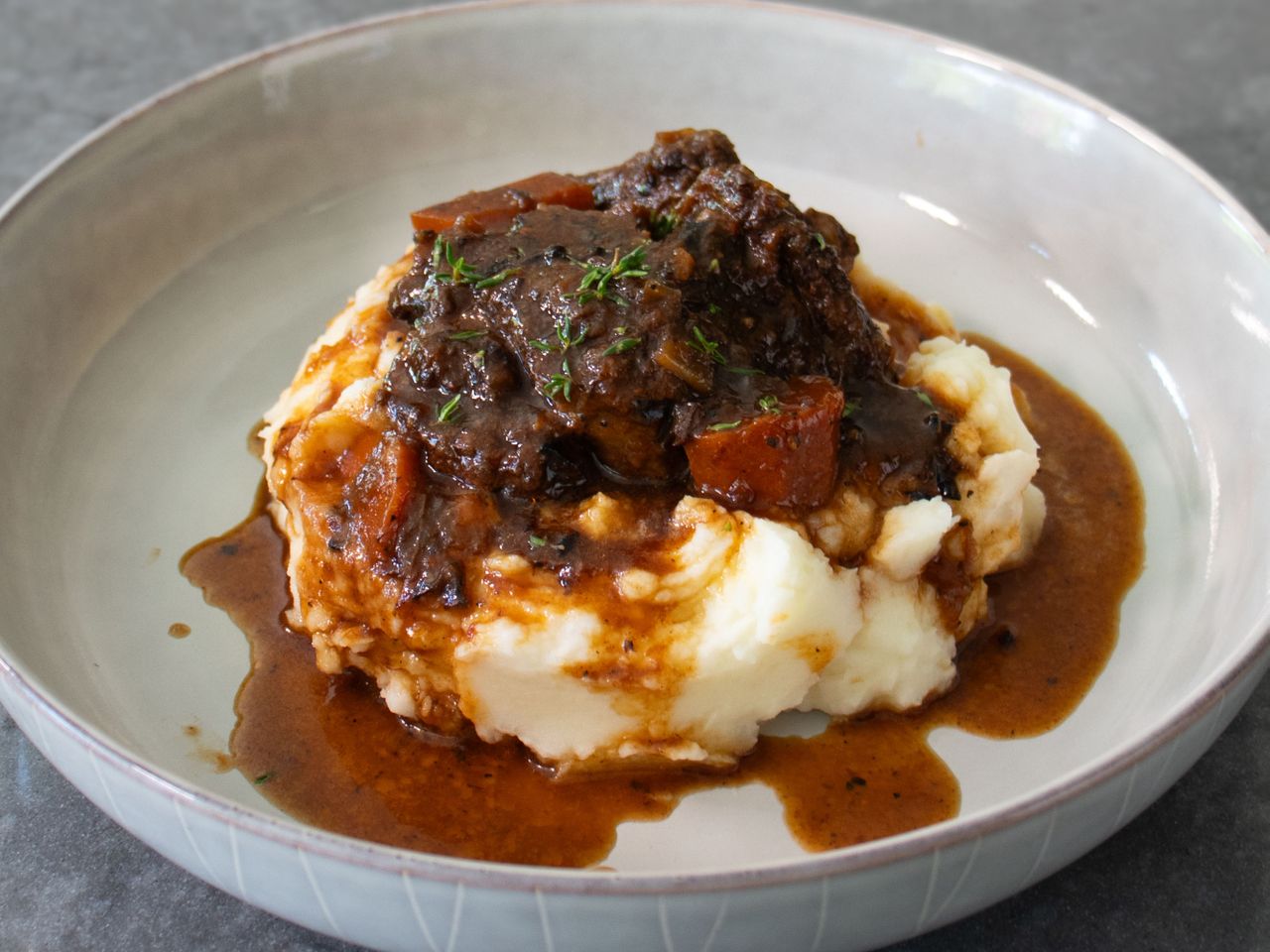closeup of a serving of short ribs with carrot chunks and a rich-looking sauce served over mashed potatoes and garnished with fresh thyme