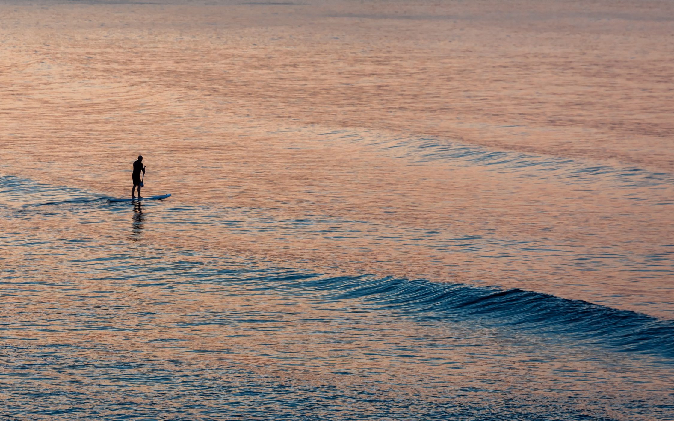 Scarborough, Yorkshire, Uk. August 21, 2018. A lone boarder stands on his tiny little floatation device, lost among the vast sea. The sea is calm, caring and allows the sport to proceed but the vastness reminds us that the sea can change at any time. This is a time of evening twilight and we're seeing the dying colours of the evening as the sun sets.