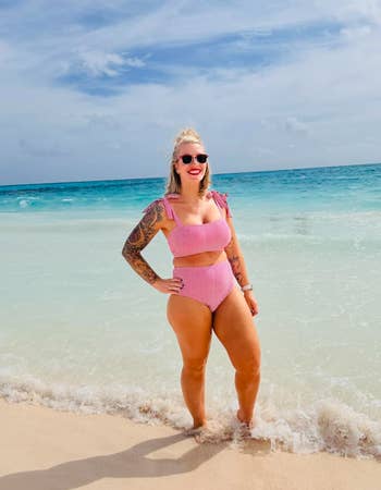 Person in beachwear stands by the ocean's edge, smiling. The image suggests a relaxed beach scene, suitable for swimwear shopping content