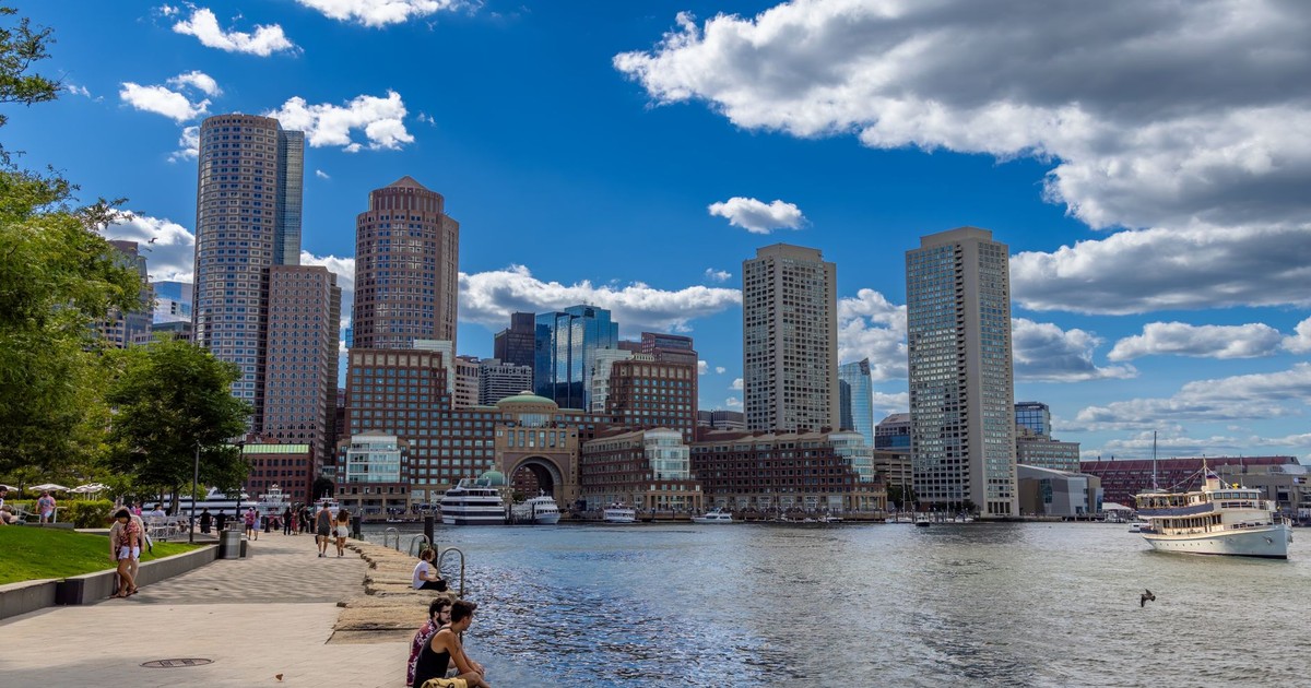 Explore the vibrant Boston harbor front with towering skyscrapers and clear blue skies reflecting on the water.
