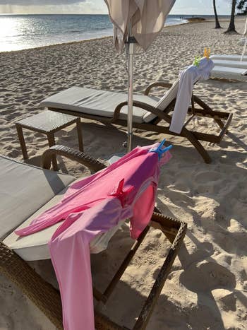 Two lounge chairs on a beach with sun umbrellas. Pink swimwear is clipped to one chair, suggesting a relaxed, sunny vacation setting
