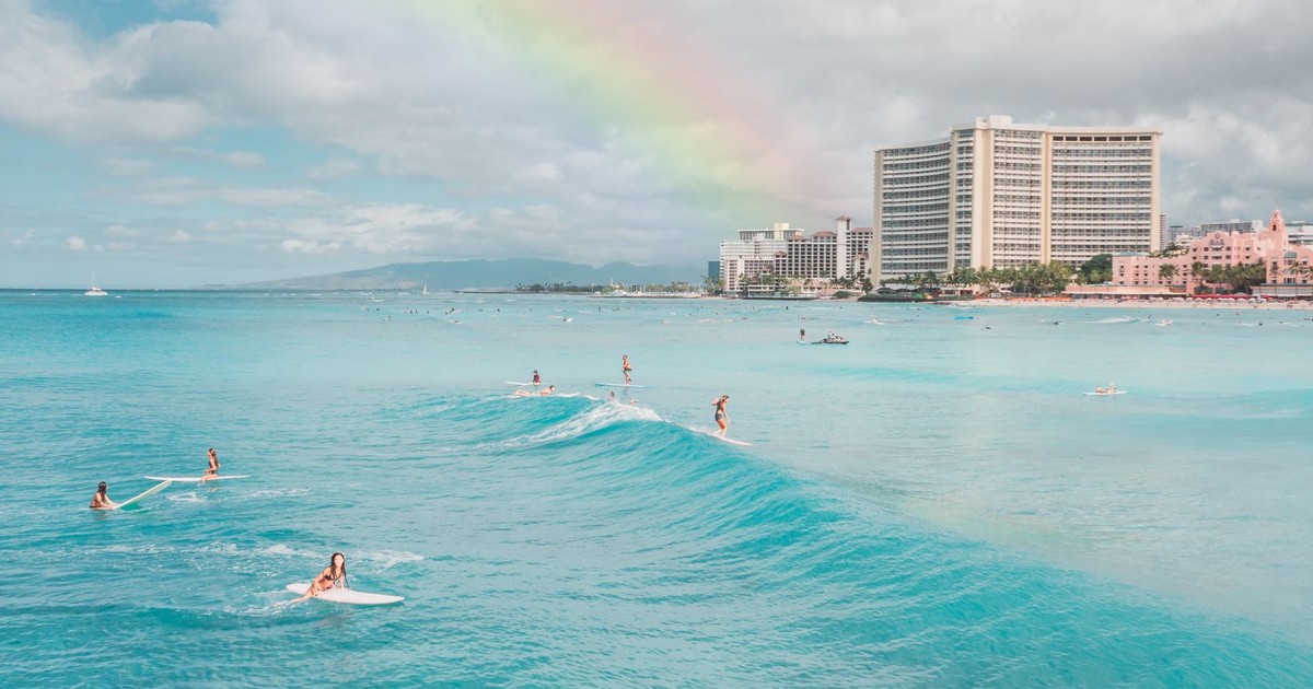 Surfers ride gentle waves beneath a vibrant rainbow at Waikiki Beach with hotels in the background.
