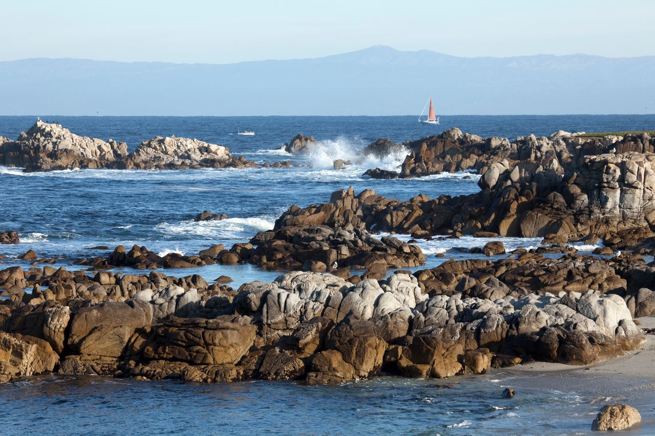 Rocky coastal scene with waves splashing and a sailboat in the distance