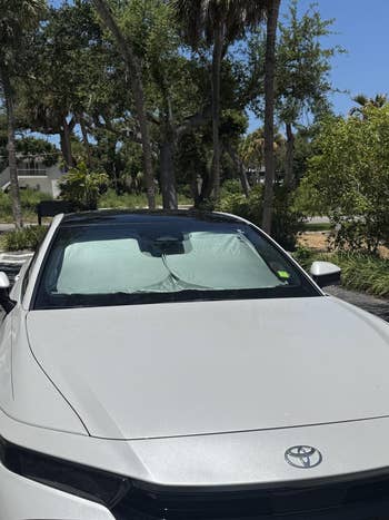 Car with sunshade in windshield parked under trees, providing UV protection and interior cooling benefits. Suitable for summer outdoors