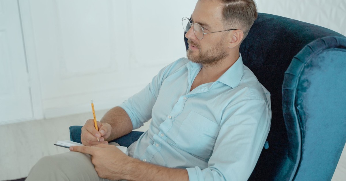A therapist in a session, wearing glasses and taking notes while seated on an armchair.