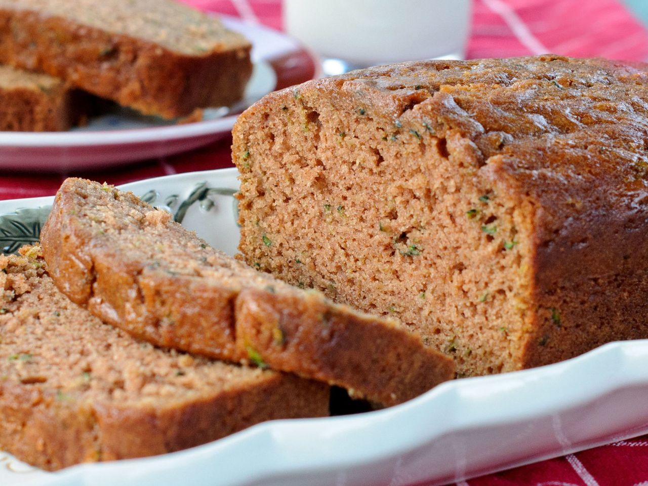 side view of a loaf of zucchini bread, partially sliced, on a white ceramic dish with a glass of milk and plate of two slices in the background