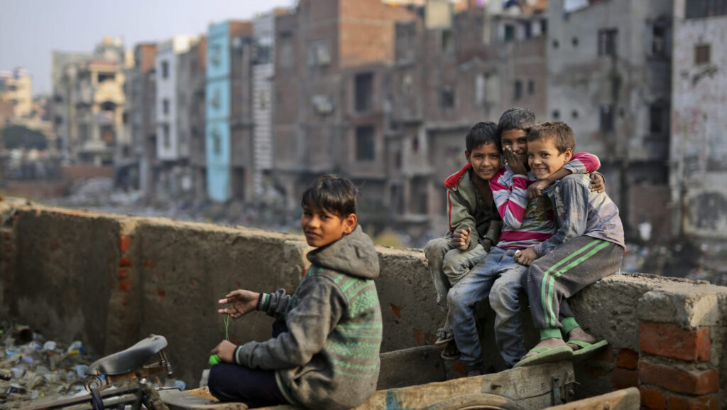 Four children sitting and playing on a concrete wall in an urban slum neighborhood, smiling at the camera