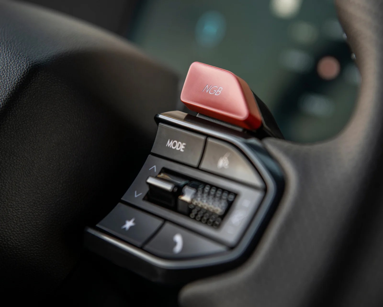 Close-up of a car steering wheel control panel with a red paddle shifter labeled "NGB" and buttons including "MODE" and phone icons.