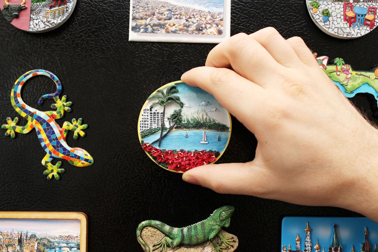 Hand placing a round travelthemed magnet on a refrigerator with other magnets visible around it