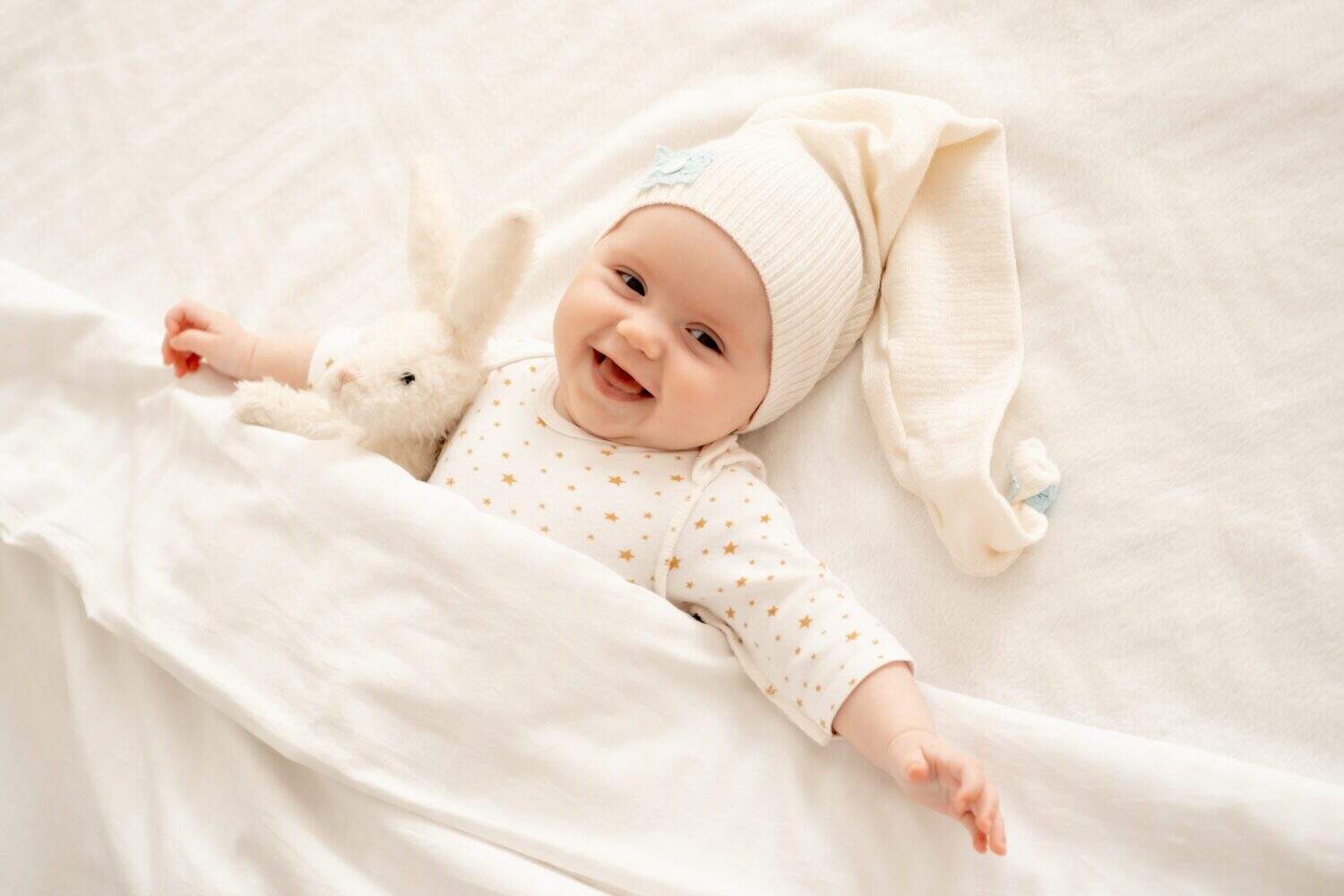 a happy newborn baby before going to bed in a sleeping cap under a blanket lies on the back and smiles on the white bed in the bedroom, a happy cute little baby, top view.