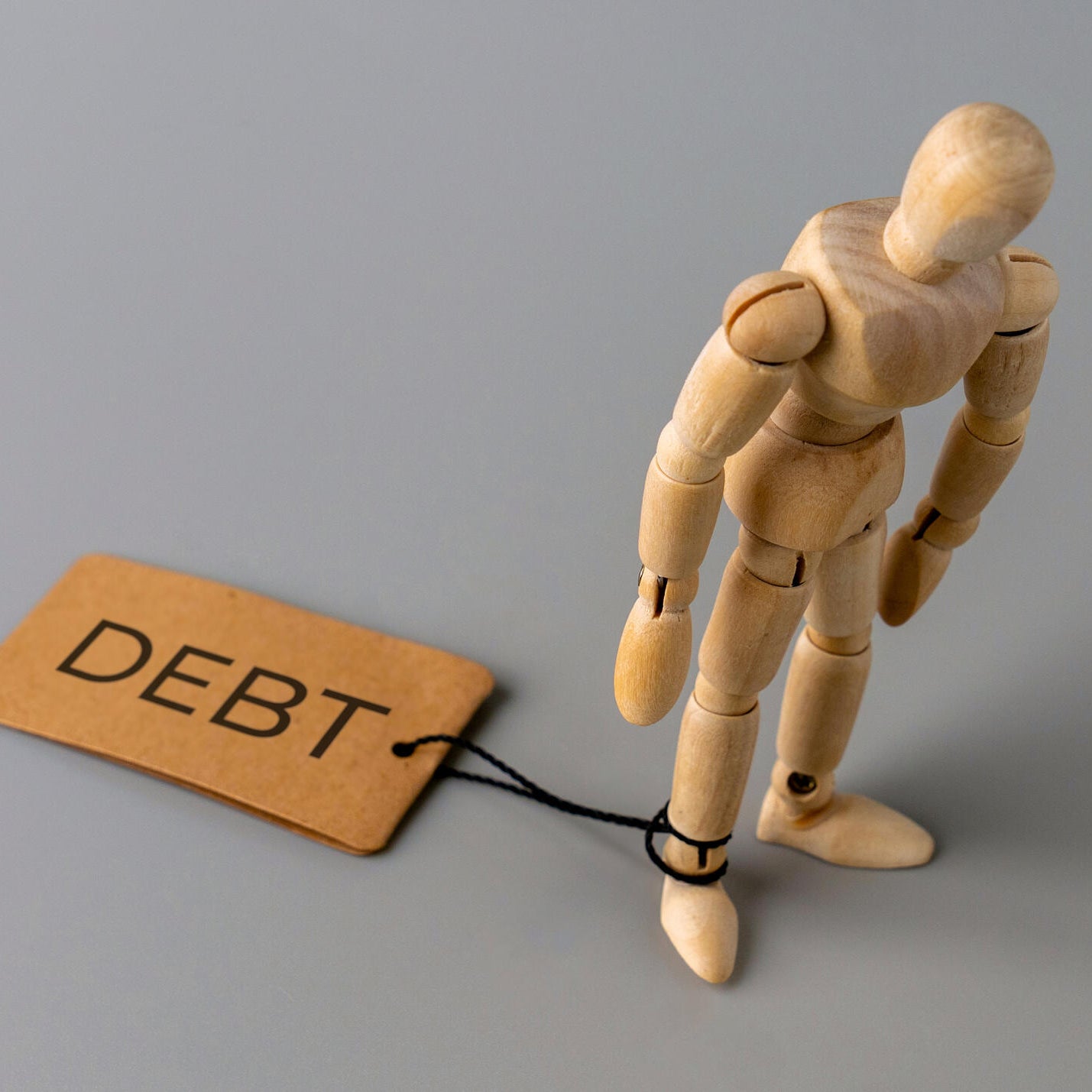 A wooden figure stands with one ankle tied by a string to a tag labeled DEBT.