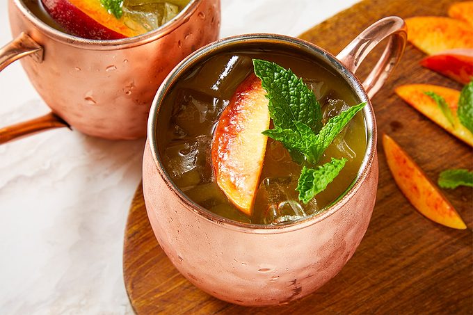 Eye Level horizontal image of two copper mugs filled with an iced drink, garnished with a peach slice and fresh mint sprigs. Additional peach slices and mint leaves are scattered on a wooden board next to the mugs, set against a white marble-like surface.
