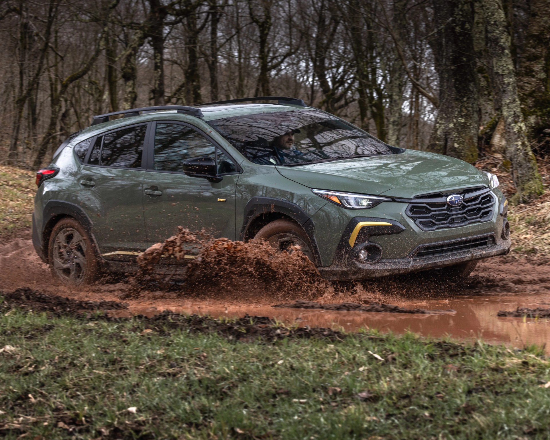 Green Subaru SUV splashing mud while driving off-road in a forested area.