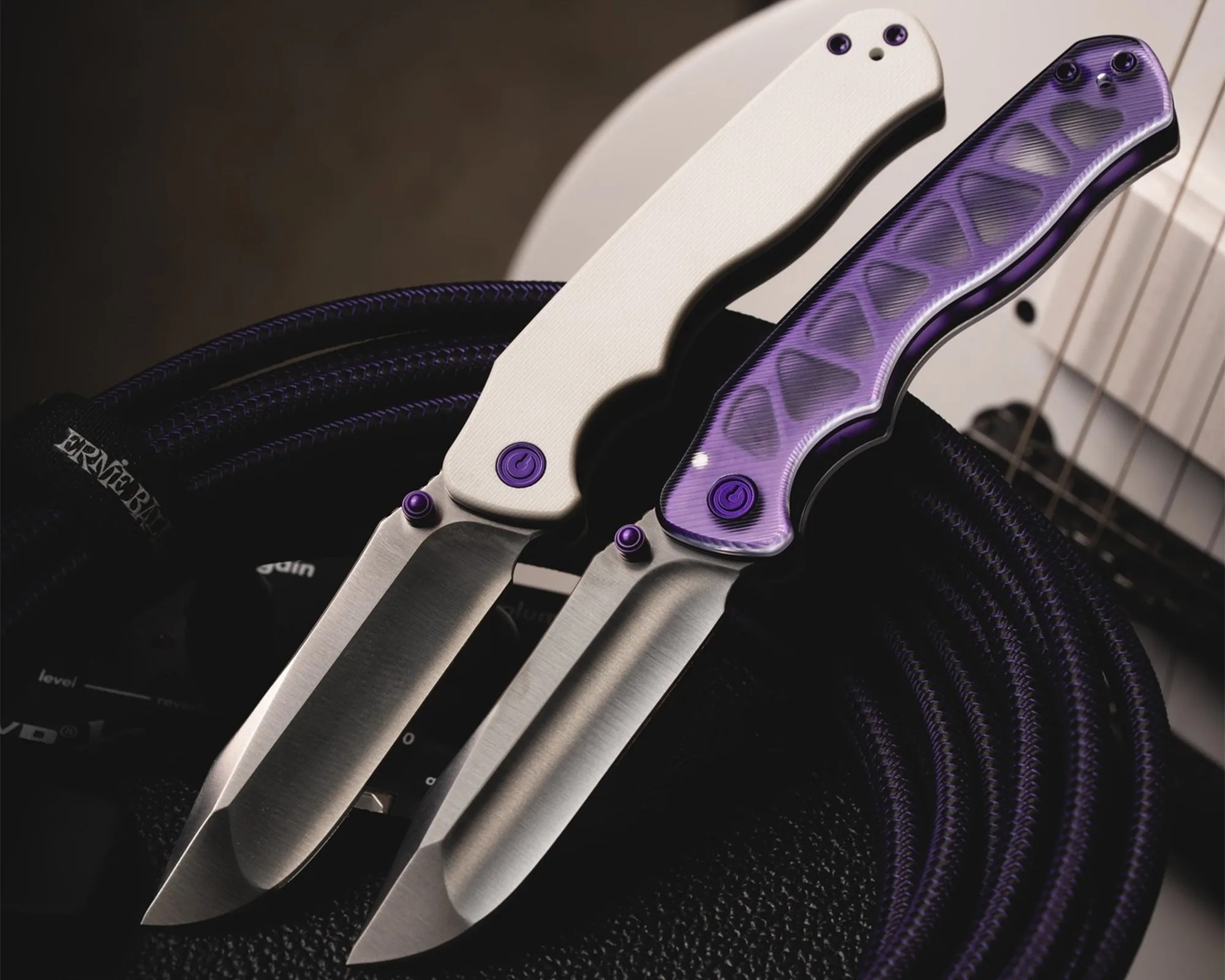Two folding knives with stainless steel blades and textured handles, one white and one translucent purple, resting on a coiled purple rope.