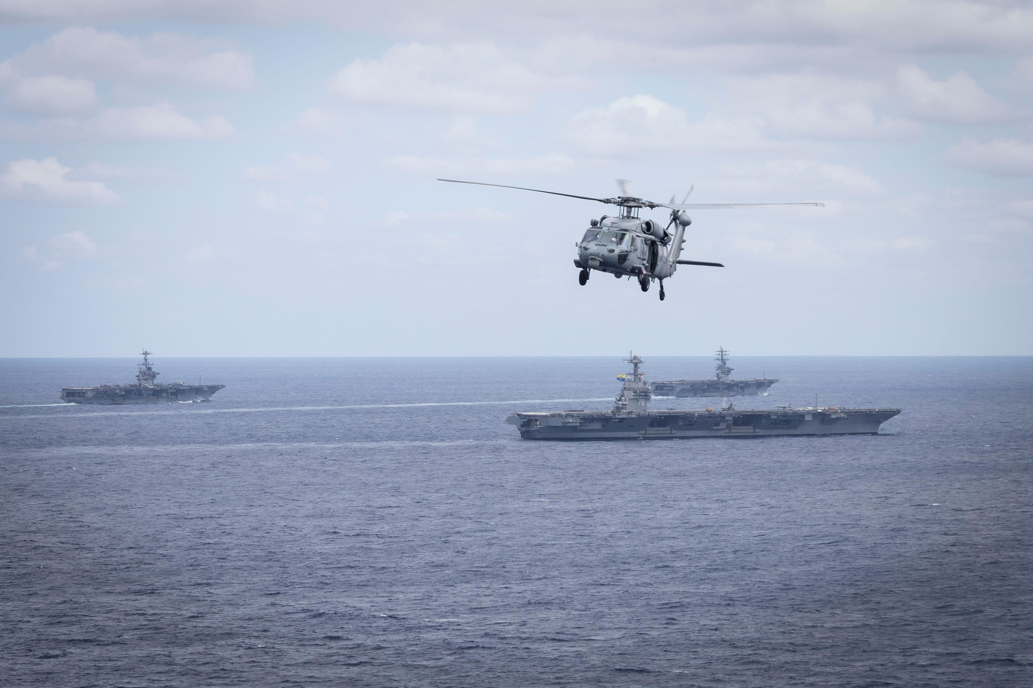 Three aircraft carriers sail in dark blue waters as a helicopter flies above in a cloudy blue sky.
