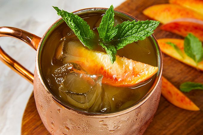 Overhead horizontal image of a cold beverage served in a copper mug. The drink contains ice cubes, a peach slice, and a fresh mint sprig garnish. Additional peach slices are visible on a wooden surface in the background.