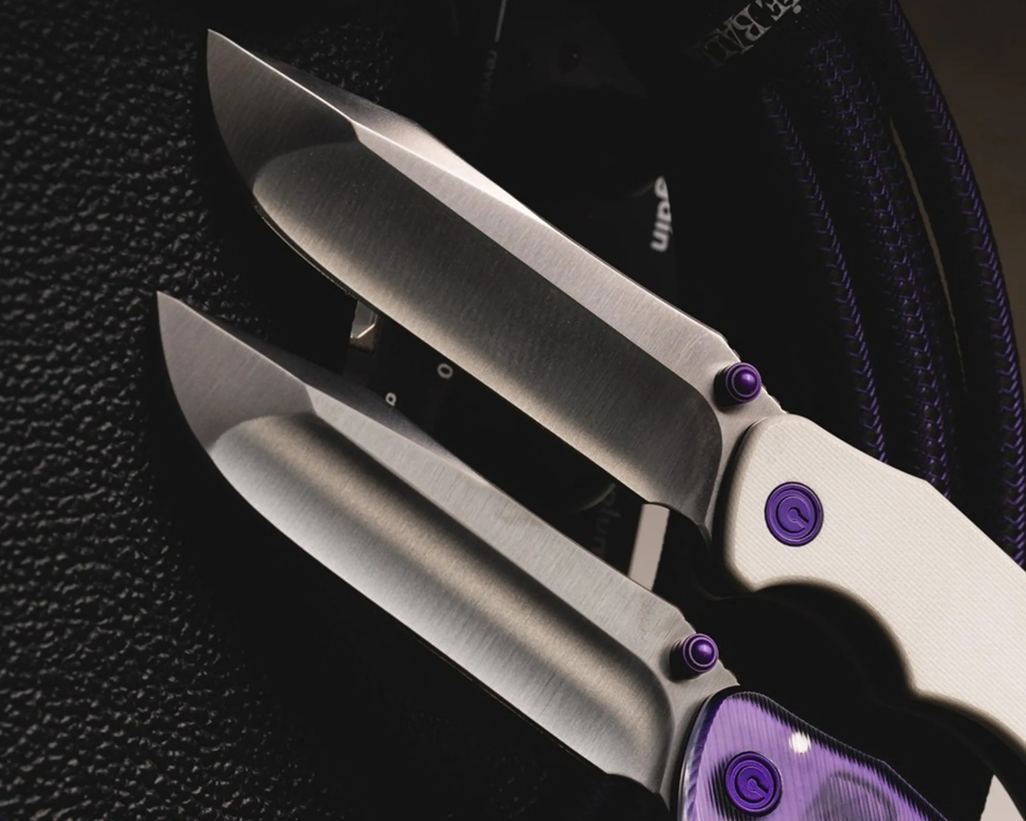 Three stainless steel folding knives with purple accents on a black textured surface.