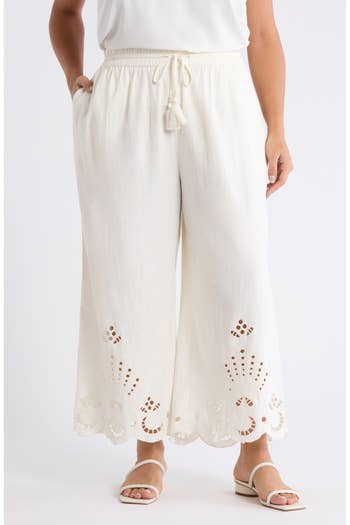 White wide-leg pants with a high waist, drawstring, and embroidered cut-out details at the hem. Paired with flat sandals