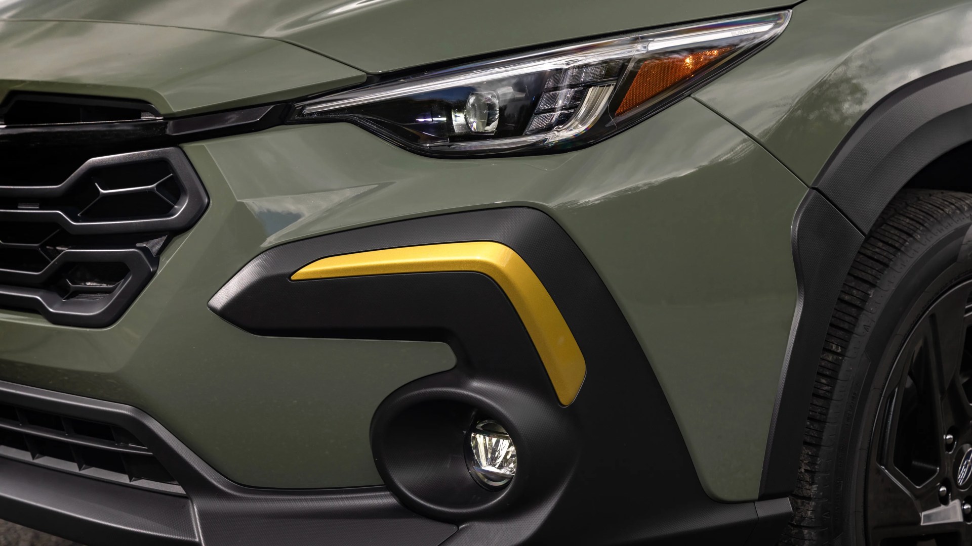 Close-up of a green car front with black grille, angular headlight, yellow accent on black bumper, and fog light.