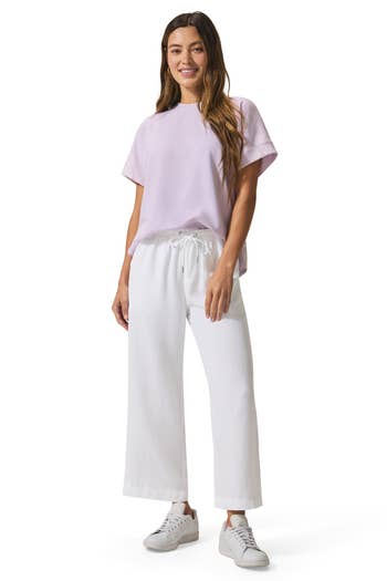 Woman wearing a casual lavender tee with white drawstring wide-leg pants and white sneakers, standing and smiling