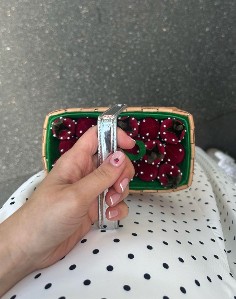 A hand holding a compact basket filled with fruit-shaped push pins on a dotted fabric background