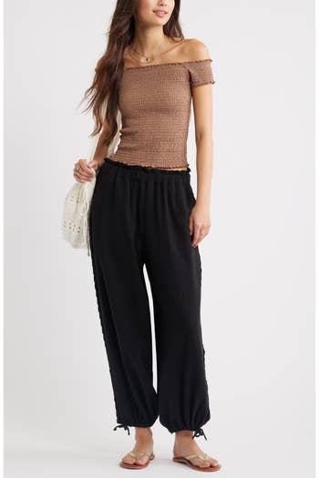 A person models a casual outfit with an off-the-shoulder top, loose pants, a crocheted bag, and sandals