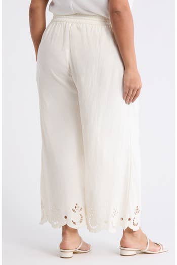 A person is wearing stylish, wide-legged white pants with embroidered hem detailing, paired with white sandals