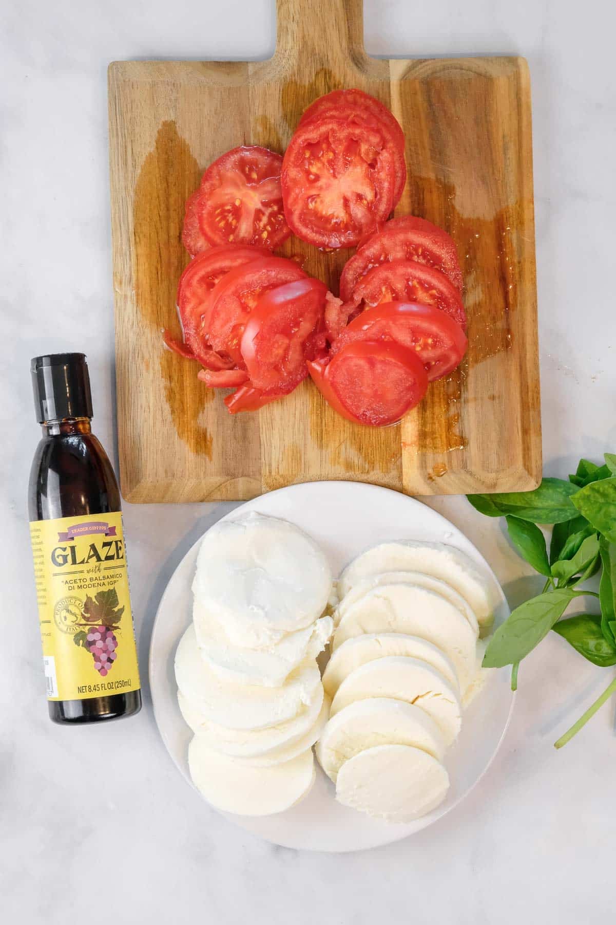 Sliced tomatoes on a wooden board, sliced mozzarella on a plate, basil leaves, and a bottle of balsamic glaze on a white surface.