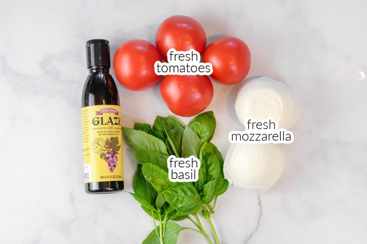 A bottle of balsamic glaze, four tomatoes, a bunch of fresh basil, and two balls of fresh mozzarella are arranged on a white surface.