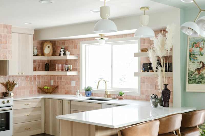 Modern kitchen with pink tiled backsplash, white countertops, wooden shelves, and decorative plants.