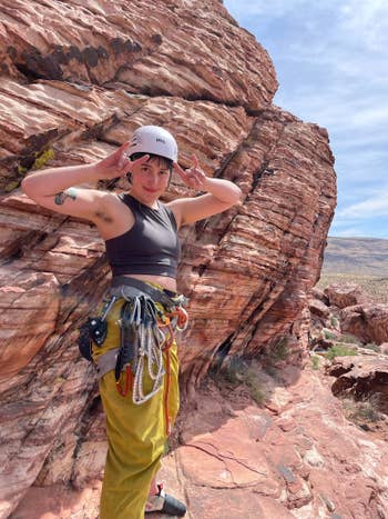 Person rock climbing, wearing a helmet, crop top, and climbing gear. Rocky landscape in the background