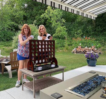 Two women enjoying a game of oversized Connect Four in a backyard setting, surrounded by greenery and outdoor furniture