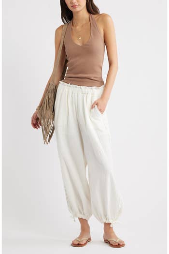 Person in a sleeveless top, loose white pants, sandals, and holding a fringed bag, showcasing a casual outfit for a shopping article