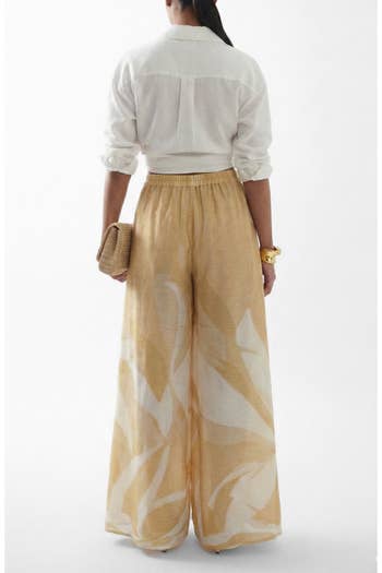 Person in a white shirt and flowing wide-leg pants holding a woven clutch, viewed from behind