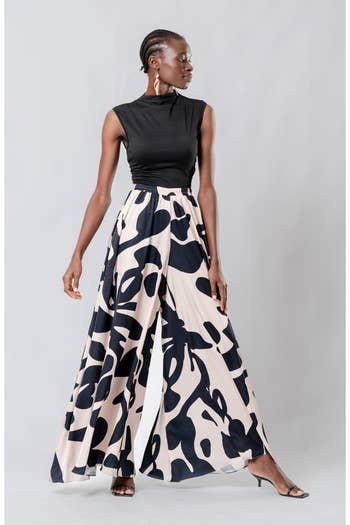 Model wearing sleeveless black top with wide-leg patterned pants and drop earrings, posing confidently for fashion shopping feature