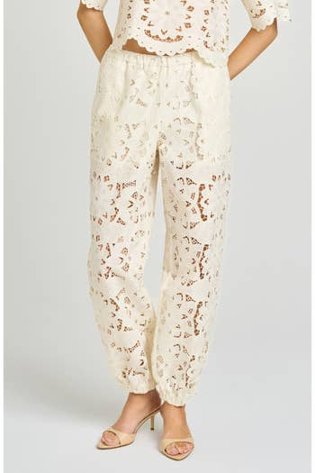 White lace pants with an intricate floral pattern, paired with matching top and beige heels. Fashion-forward style for shopping article