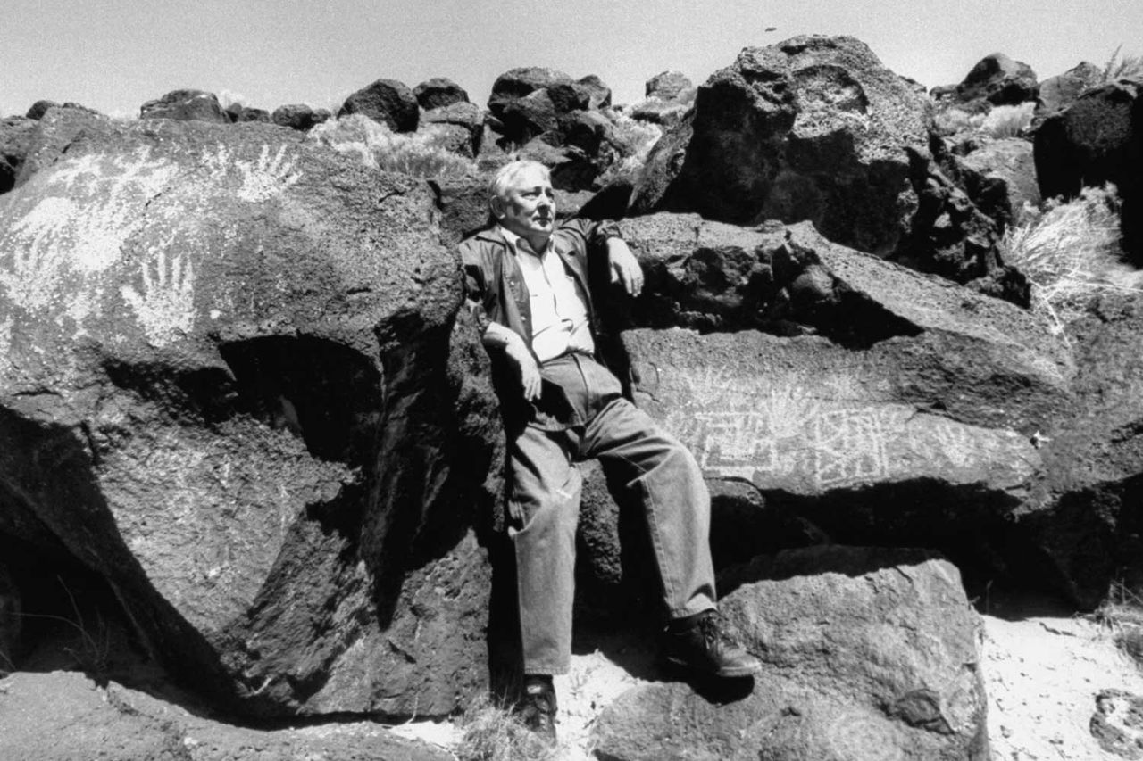 Writer Tony Hillerman standing amongst rocks where prehistoric Navajo rock drawings are apparent.