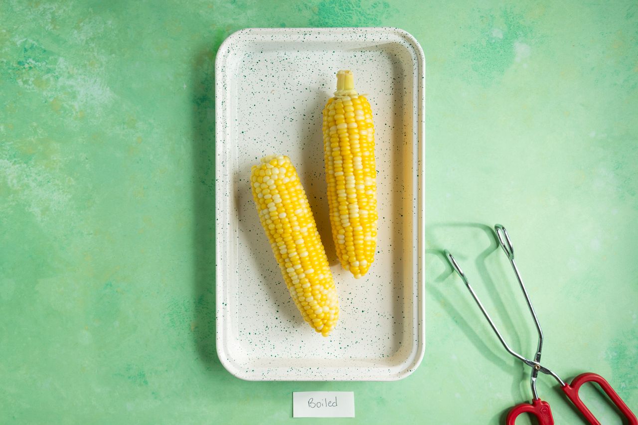 Two boiled corn cobs on a tray with tongs placed next to it on a green surface