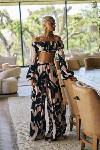 A person models a stylish off-shoulder top and wide-leg pants with an abstract pattern in a modern living room setting