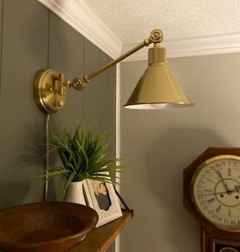 Brass wall-mounted lamp extended over a wooden bowl, with a potted plant and a photo on a table next to a clock