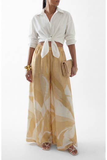 Person in a tied white shirt, flowy wide-leg pants, holding a woven clutch and wearing strappy heels