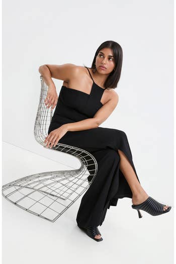 Woman in a sleek, asymmetrical black dress sits on a modern metal chair, wearing open-toe black heels