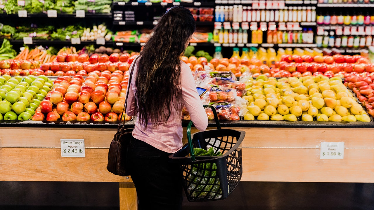 Woman with long, dark hair seen from behind checking grocery list on phone in fruit aisle of grocery store.