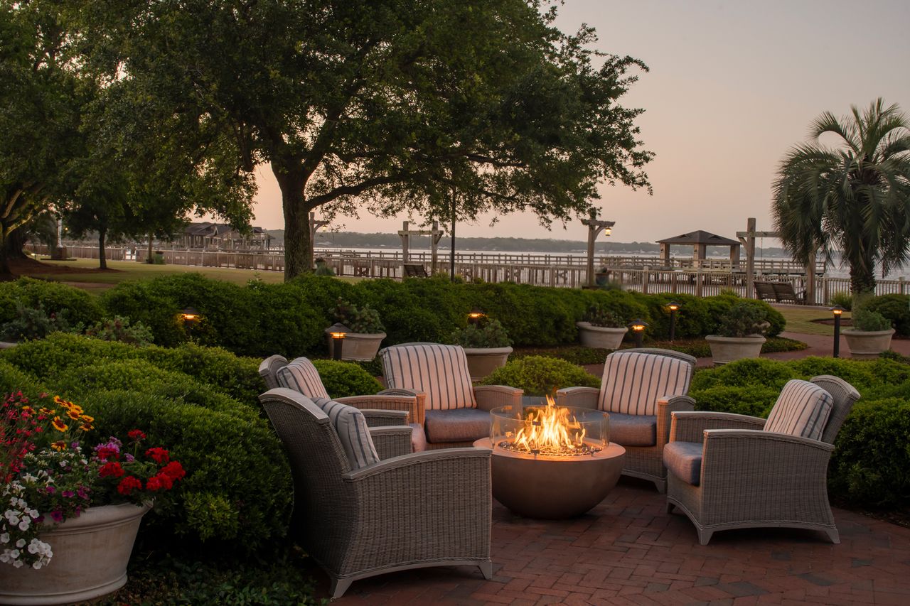 Outdoor seating area with a fire pit surrounded by greenery and a view of a dock and water in the background