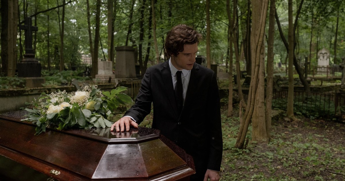 A man in a black suit mourns at a funeral in a lush green cemetery, leaning on the coffin.