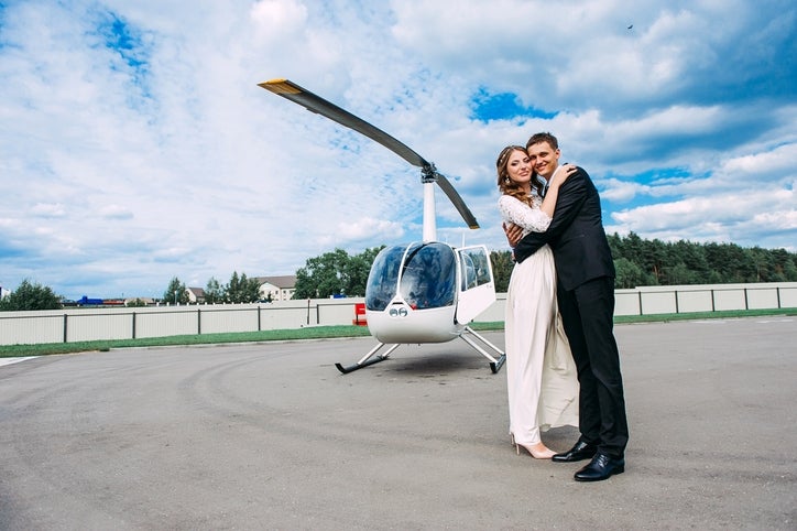 A couple in elegant attire embraces in front of a helicopter on a tarmac, suggesting a luxurious and adventurous lifestyle