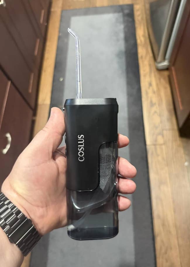 Hand holding a portable, handheld water flosser with a clear tank and a slim straw-like nozzle. Brand name