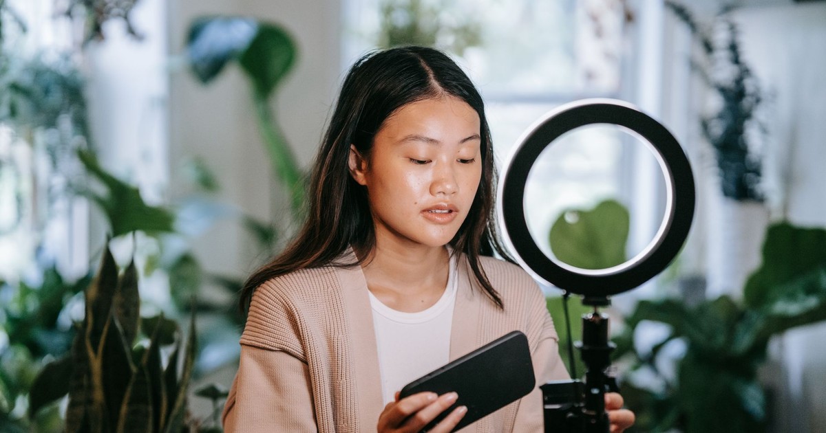 Asian woman adjusting smartphone and ring light indoors for video recording.