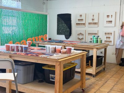 View of an artist's studio with various beads in plastic containers and works arranged on two tables. In the background is a beaded curtain that reads 'PLEASURE'.