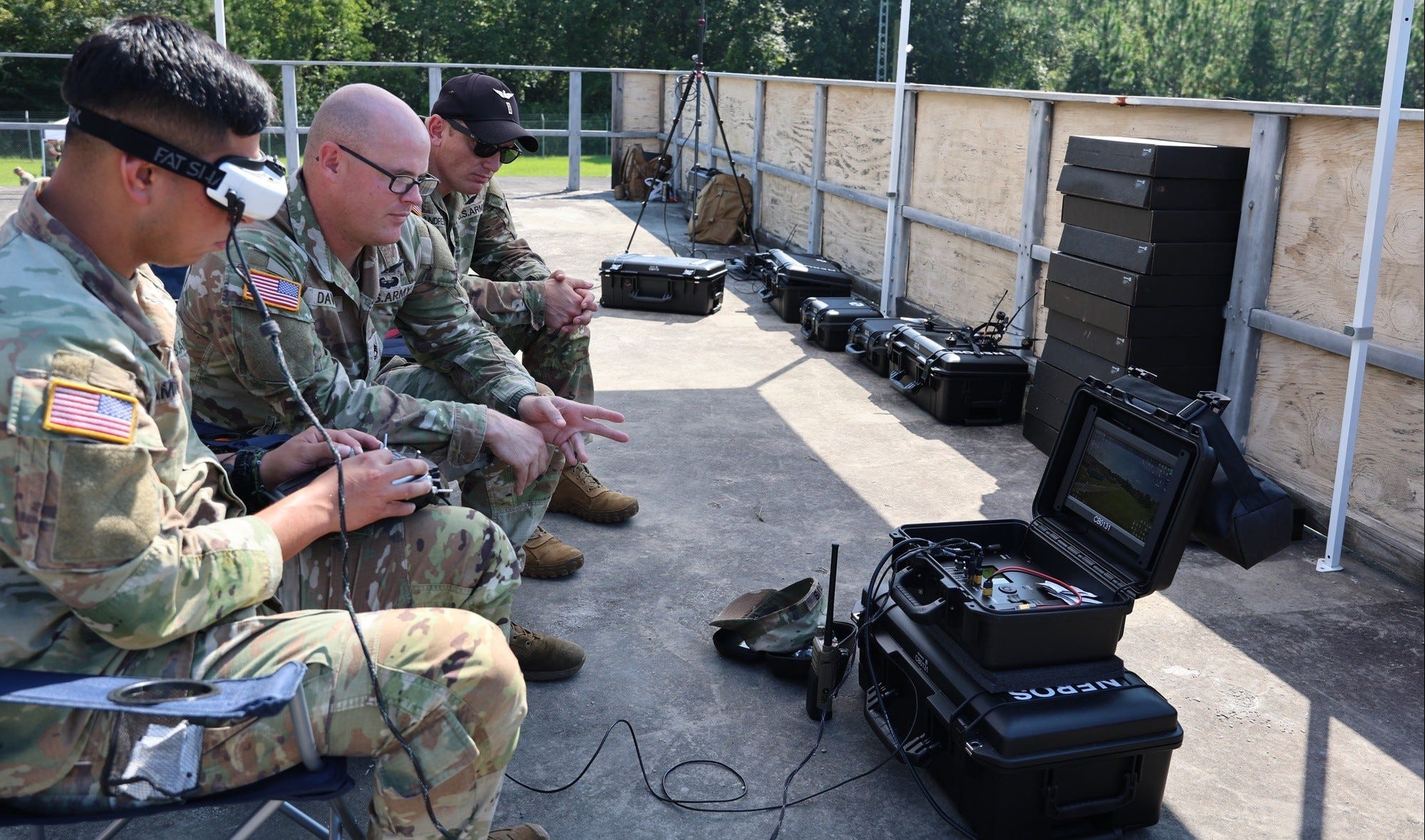 Military personnel in camouflage uniforms sit beside portable electronic equipment cases during an outdoor training setup.